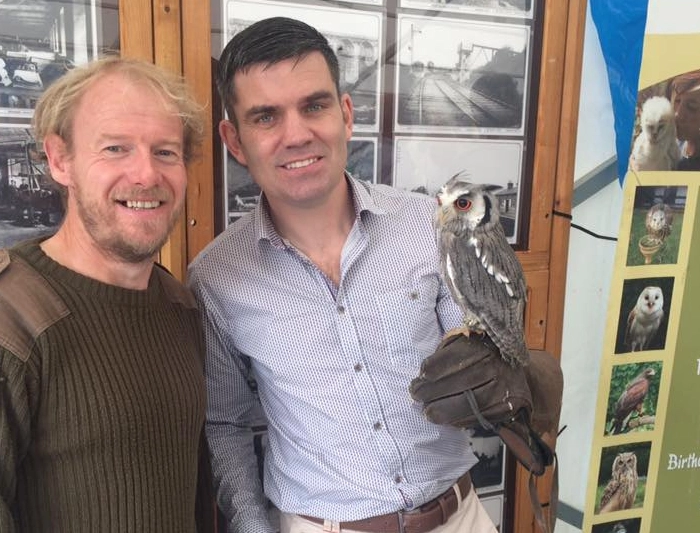 Two men smiling while one holds a small, grey-faced owl on a leather falconry glove.