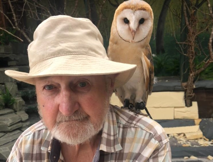 An elderly man in a sun hat posing for a photo with a barn owl perched on his shoulder.