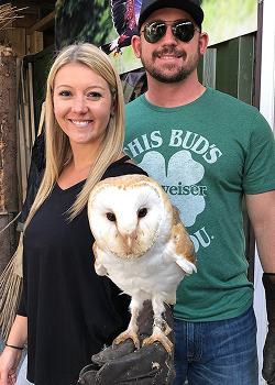 A smiling couple posing with a white Barn Owl perched on a handler's leather glove.