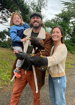 A smiling family of three posing outdoors with a Harris's hawk perched on a falconry glove.