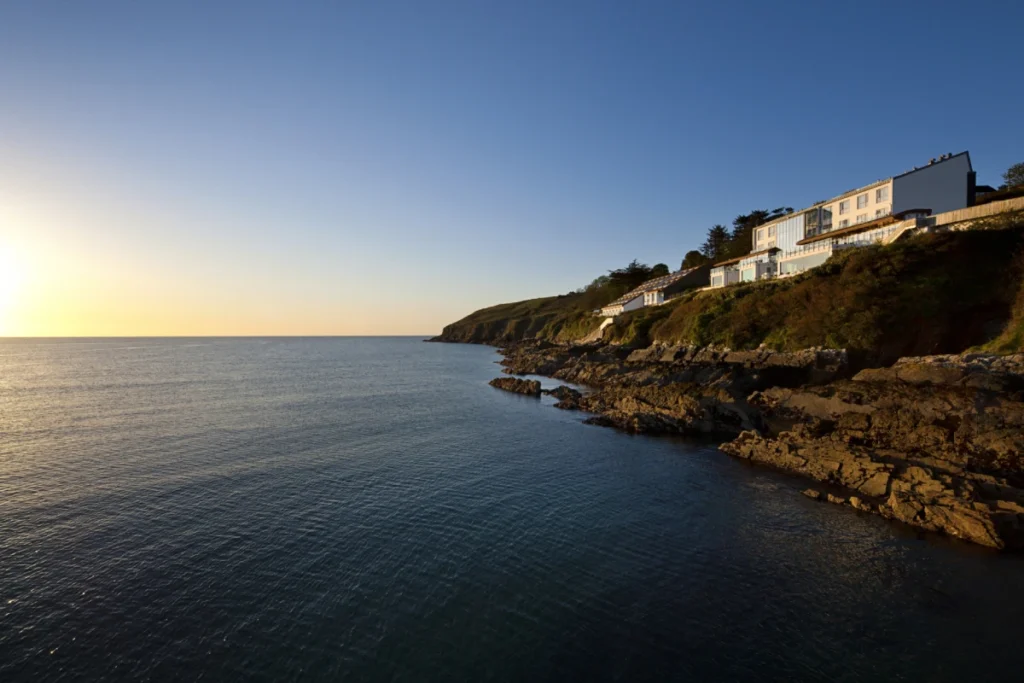 A coastal hotel sits perched on a rocky cliffside overlooking the calm blue sea at sunset.
