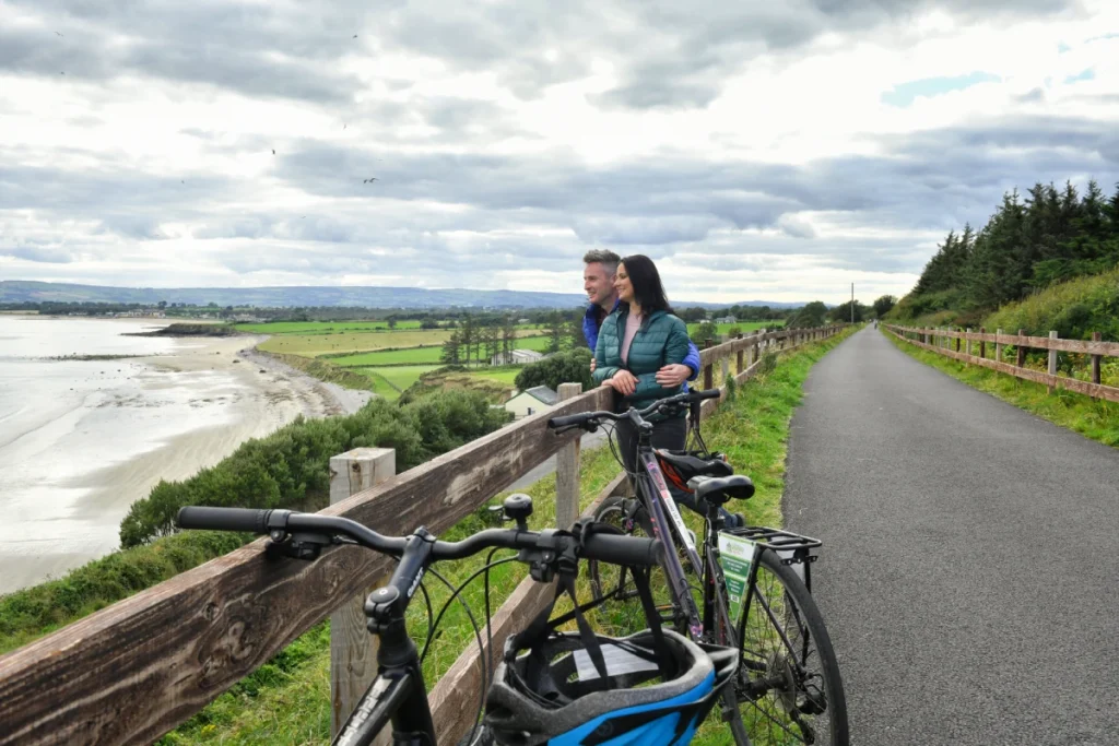 A couple stands with their bicycles along a coastal paved path, looking out over a beach and green fields under a cloudy sky.