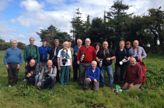 A large group of photography enthusiasts poses together in a green field, many holding professional cameras with long telephoto lenses.