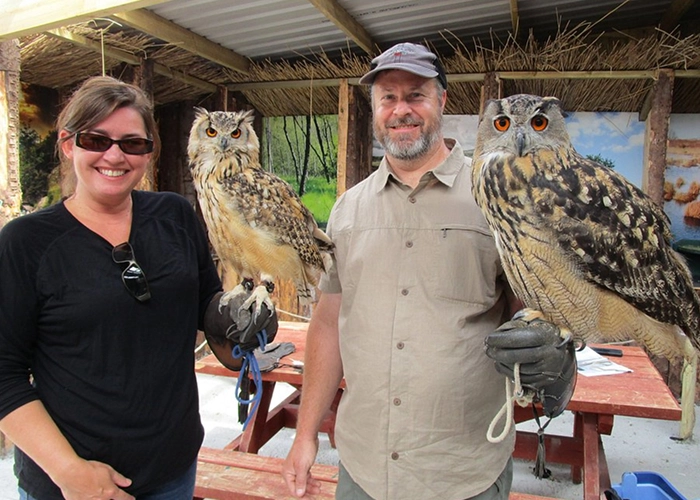 Two people smiling while holding large owls on leather gloves.